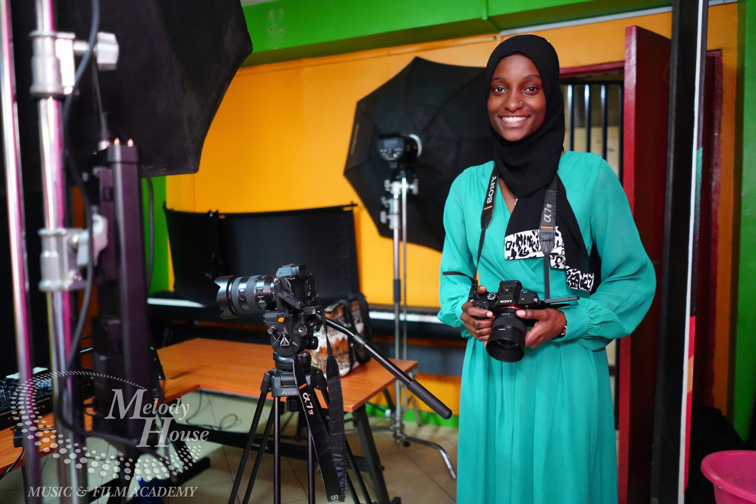 A smiling student holds a professional Sony Alpha camera while standing in a fully-equipped studio for photography and videography courses in Kenya at Music And Film Academy. The studio setup includes a second DSLR camera on a professional tripod, softbox studio lights, and a vibrant yellow and green interior. This image highlights the inclusive, hands-on training environment where students learn to master high-end digital imaging and cinematography equipment.