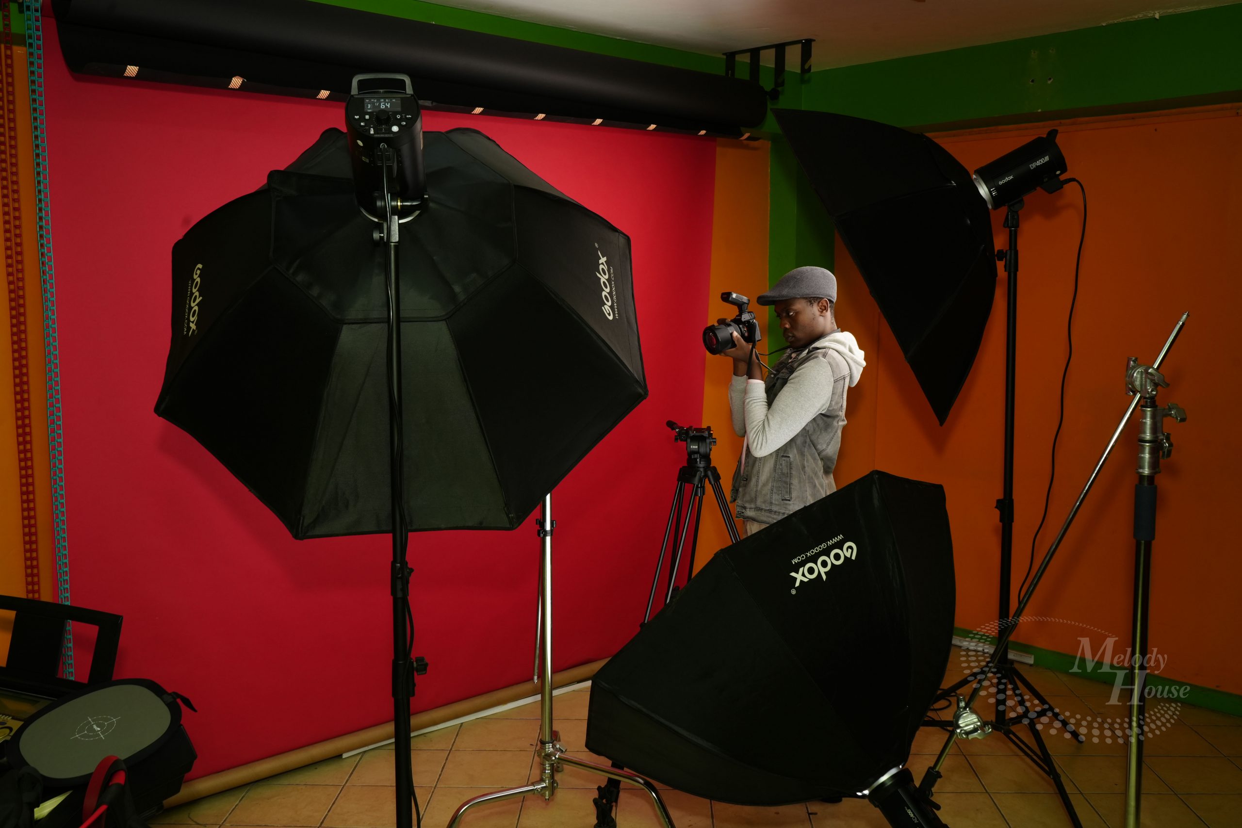 A student practices studio lighting techniques during photography courses at Music And Film Academy. The image shows a photographer behind a camera, surrounded by professional Godox lighting equipment, including large softboxes and an umbrella, set against a vibrant red and orange backdrop. This hands-on training environment illustrates the academy's focus on mastering high-end studio setups and professional cinematography tools.