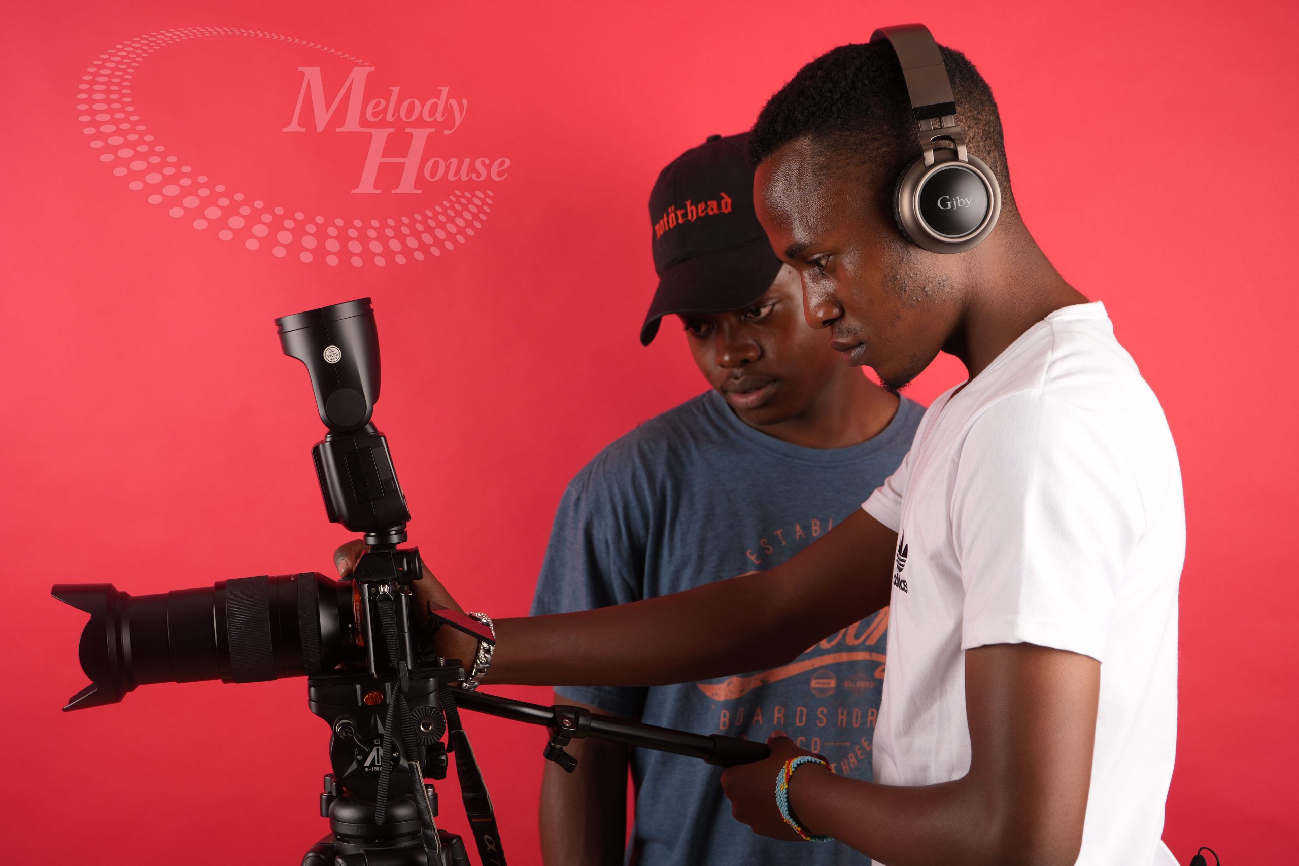 Two students collaborate on a professional camera setup during videography courses in Kenya at the Music And Film Academy. One student, wearing studio headphones, focuses on adjusting a DSLR camera mounted on a professional tripod with a fluid head, while the other student observes the technical process. Set against a vibrant red backdrop featuring the Melody House logo, the image highlights the academy's emphasis on hands-on training with industry-standard film and video equipment