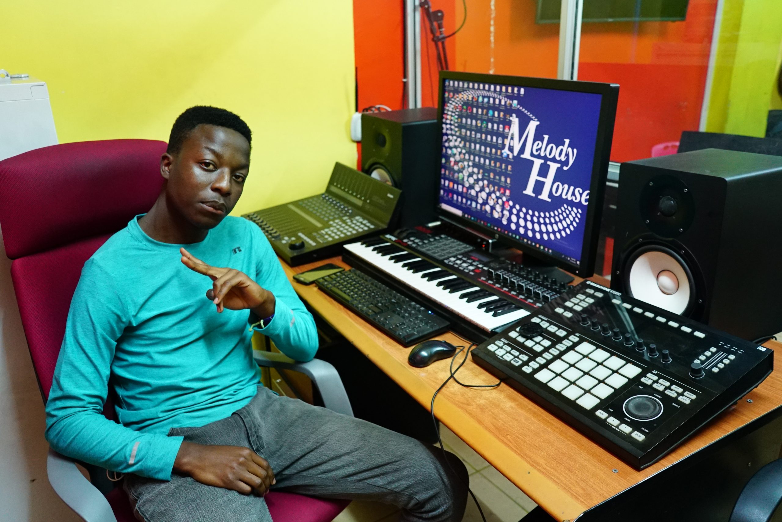 A music production student giving a peace sign at a professional workstation in Nairobi, featuring a Yamaha HS8 monitor, Native Instruments Maschine Studio, and an Akai MIDI keyboard.