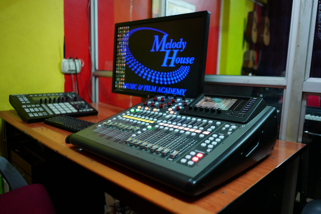 A high-tech digital audio mixing console and beat pad sit on a studio desk at Music & Film Academy. The computer monitor in the background displays the academy's logo, highlighting the professional-grade equipment used in their sound engineering courses. The vibrant studio setting includes acoustic guitars visible through a glass partition, illustrating a comprehensive creative learning environment.