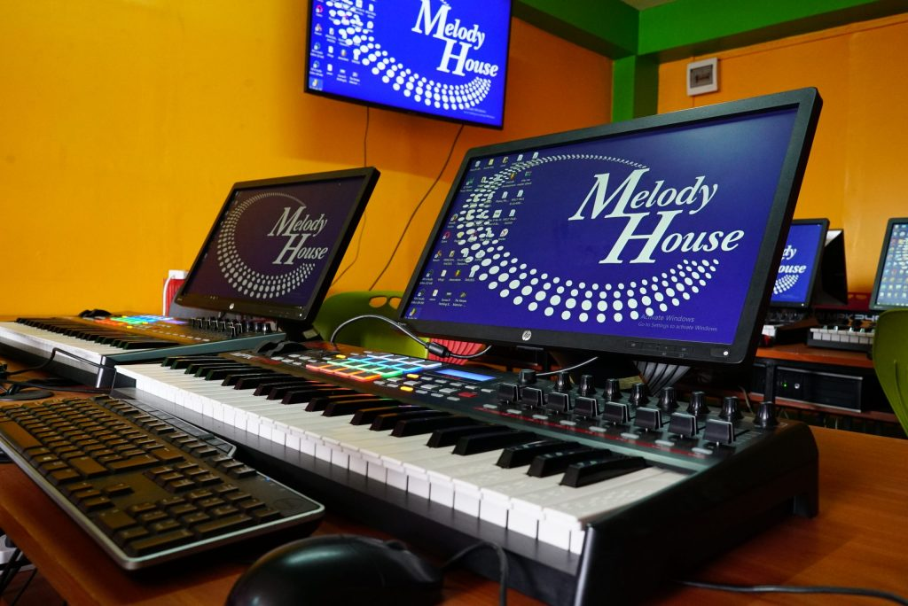 A row of professional workstations for **music production courses** at **Music And Film Academy**. Each station features an Akai MPK249 MIDI keyboard controller with colorful backlit pads, a high-resolution HP monitor displaying the Melody House logo, and a dedicated computer setup. The vibrant yellow and orange studio environment showcases the modern, hands-on learning lab where students master beat-making, sound design, and digital audio workstation (DAW) techniques.