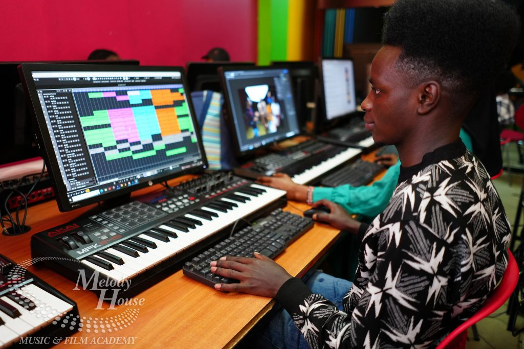 A student actively engages in music production courses at Music And Film Academy, working on a professional digital audio workstation (DAW). The student is using an Akai MPK249 MIDI keyboard to arrange colorful audio tracks and patterns on a large computer monitor. The modern classroom setup features multiple workstations, highlighting the academy’s commitment to providing students with hands-on technical training in beat-making and sound engineering.