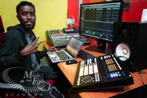 A Music and Film Academy student smiling and giving a peace sign while using a Native Instruments Maschine Studio and a digital mixing console in the Nairobi production labs.