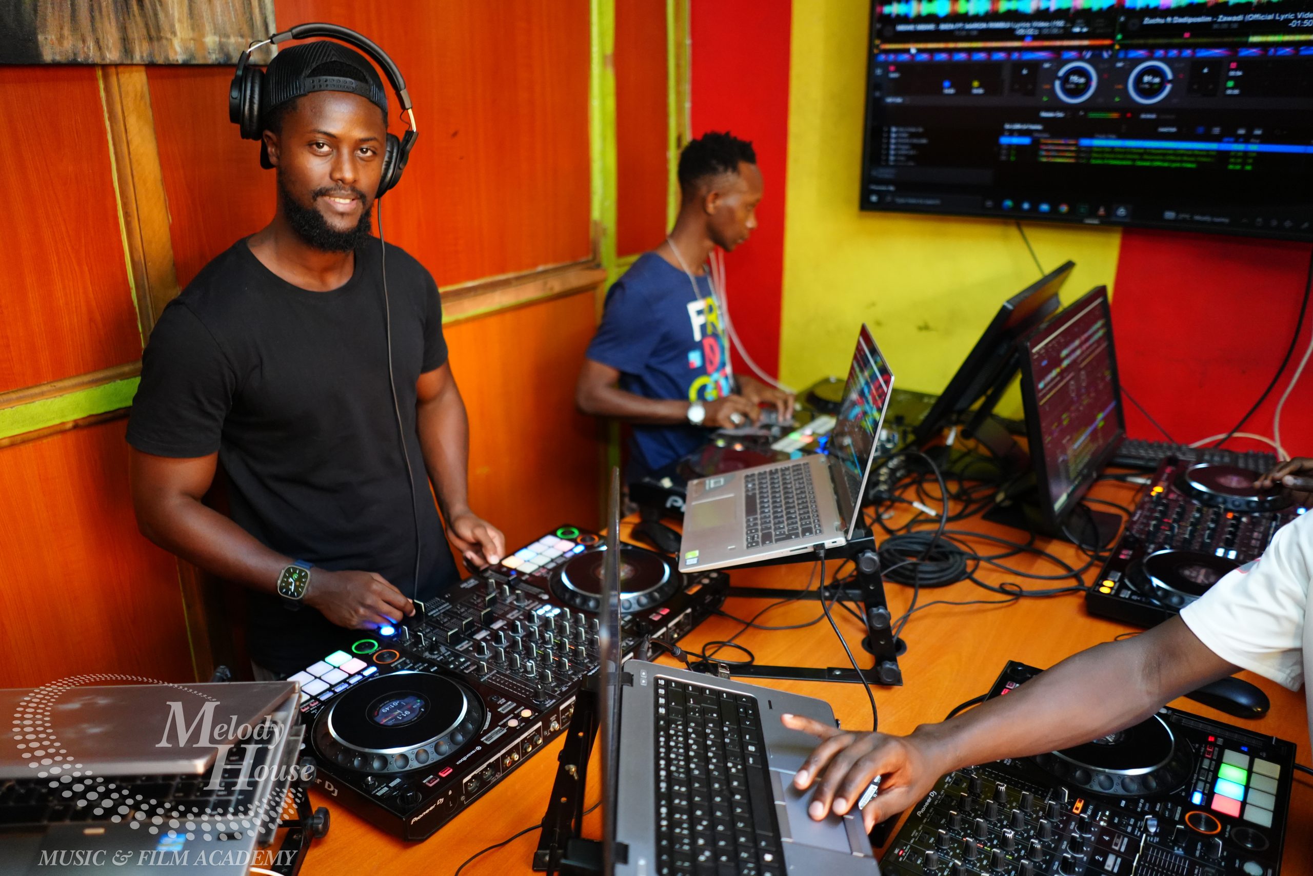 A high-angle view of two student DJs at Music And Film Academy Nairobi using Pioneer DJ controllers; one laptop displays a yellow Serato interface, and another shows a blue Rekordbox waveform screen.