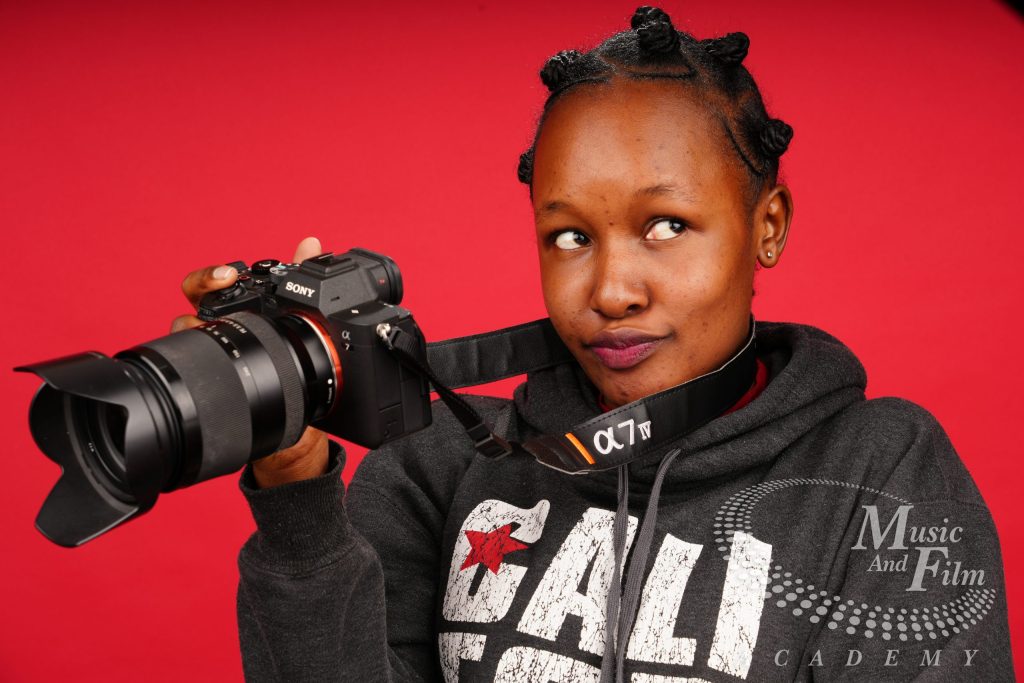 A female portrait model with deep melanin skin tones holding a professional Sony Alpha mirrorless camera against a red background during a lighting configuration lab at Music and Film Academy.
