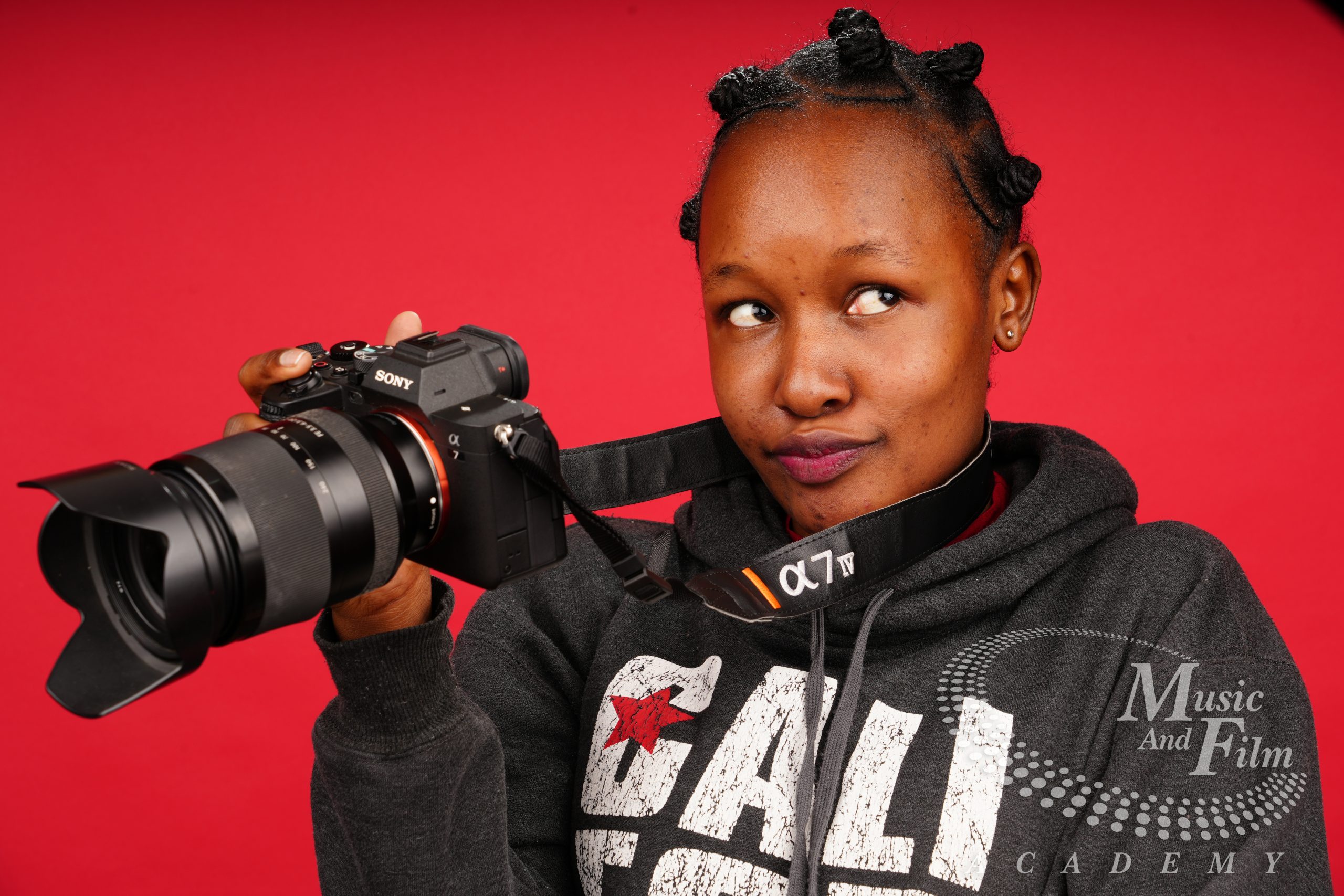 A female portrait model with deep melanin skin tones holding a professional Sony Alpha mirrorless camera against a red background during a lighting configuration lab at Music and Film Academy.