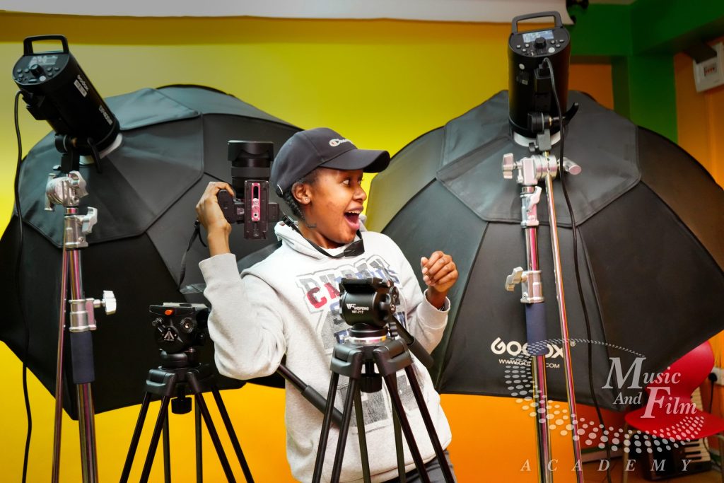 A photography student holds a DSLR camera on a tripod next to professional Godox studio strobes and large black umbrellas during an advanced certificate in photography at the Music and Film Academy in Nairobi.