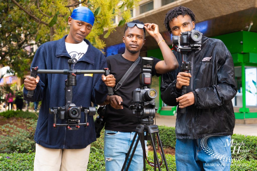 Three videography students operating a dual-handle motorized gimbal stabilizer, a camera tripod with a Godox speedlight, and a single-handed 3-axis electronic stabilizer during an outdoor practical in Nairobi.