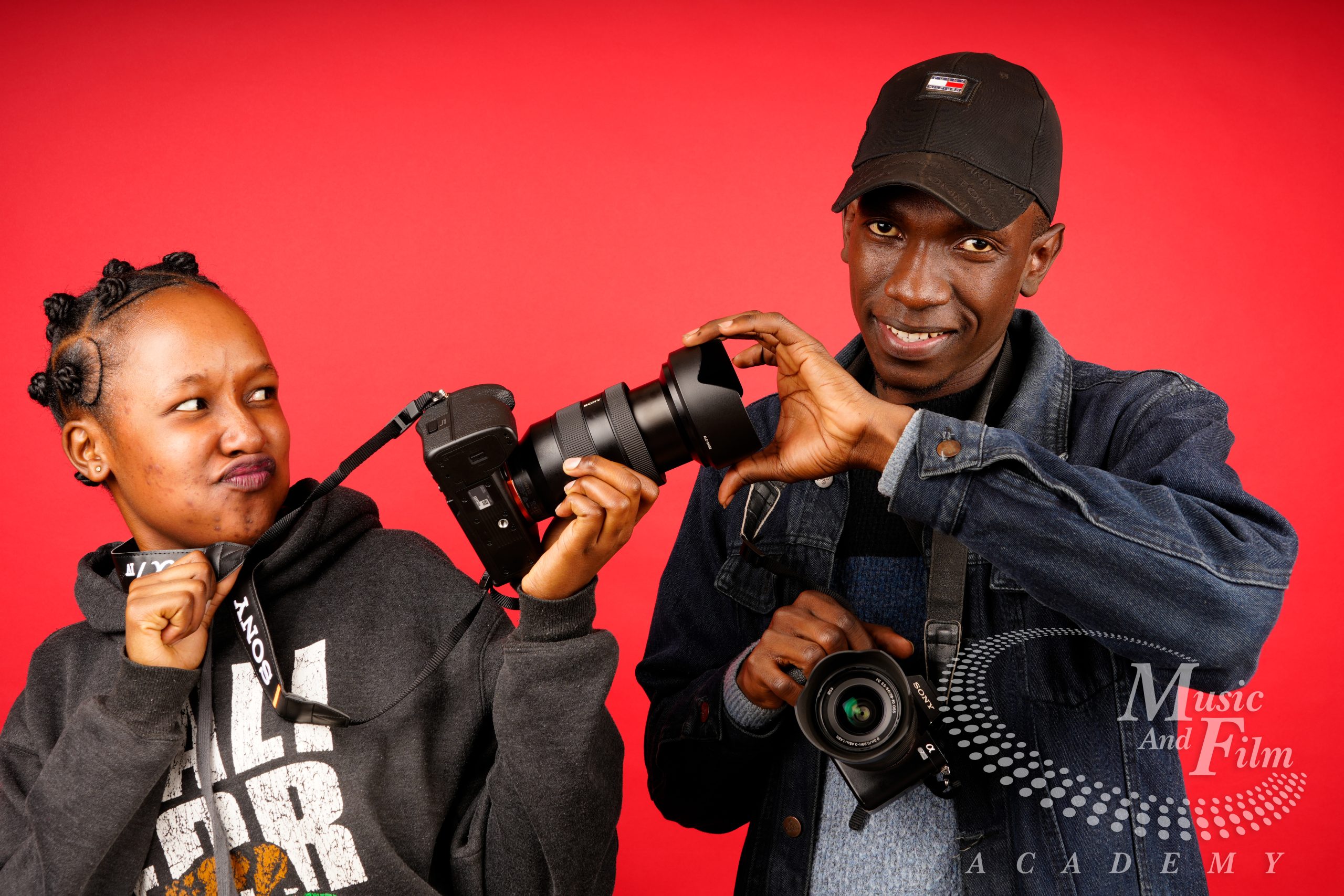 Two photography students adjusting a Sony mirrorless camera lens with a hood against a red background during a Certificate in Photography in Nairobi.