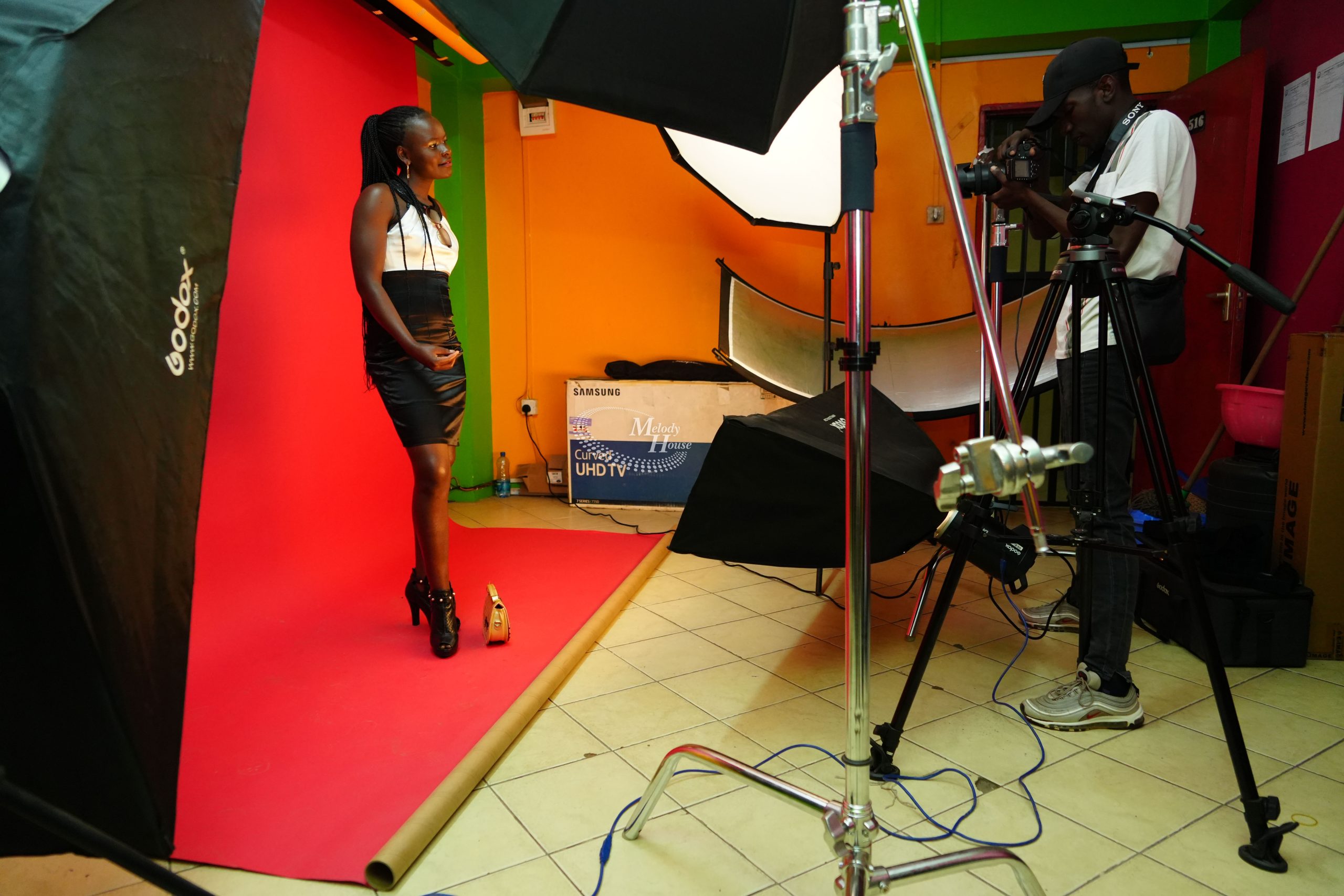 A videography student operating a camera on a professional fluid-head tripod, filming a talent standing against a red backdrop under a continuous Godox softbox lighting array in a studio in Nairobi.