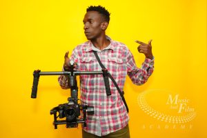 A student learning the skills for becoming a hybrid creator, operating a professional Sony mirrorless camera mounted on a dual-handle DJI Ronin gimbal stabilizer against a bright yellow background at the Music And Film Academy studio.