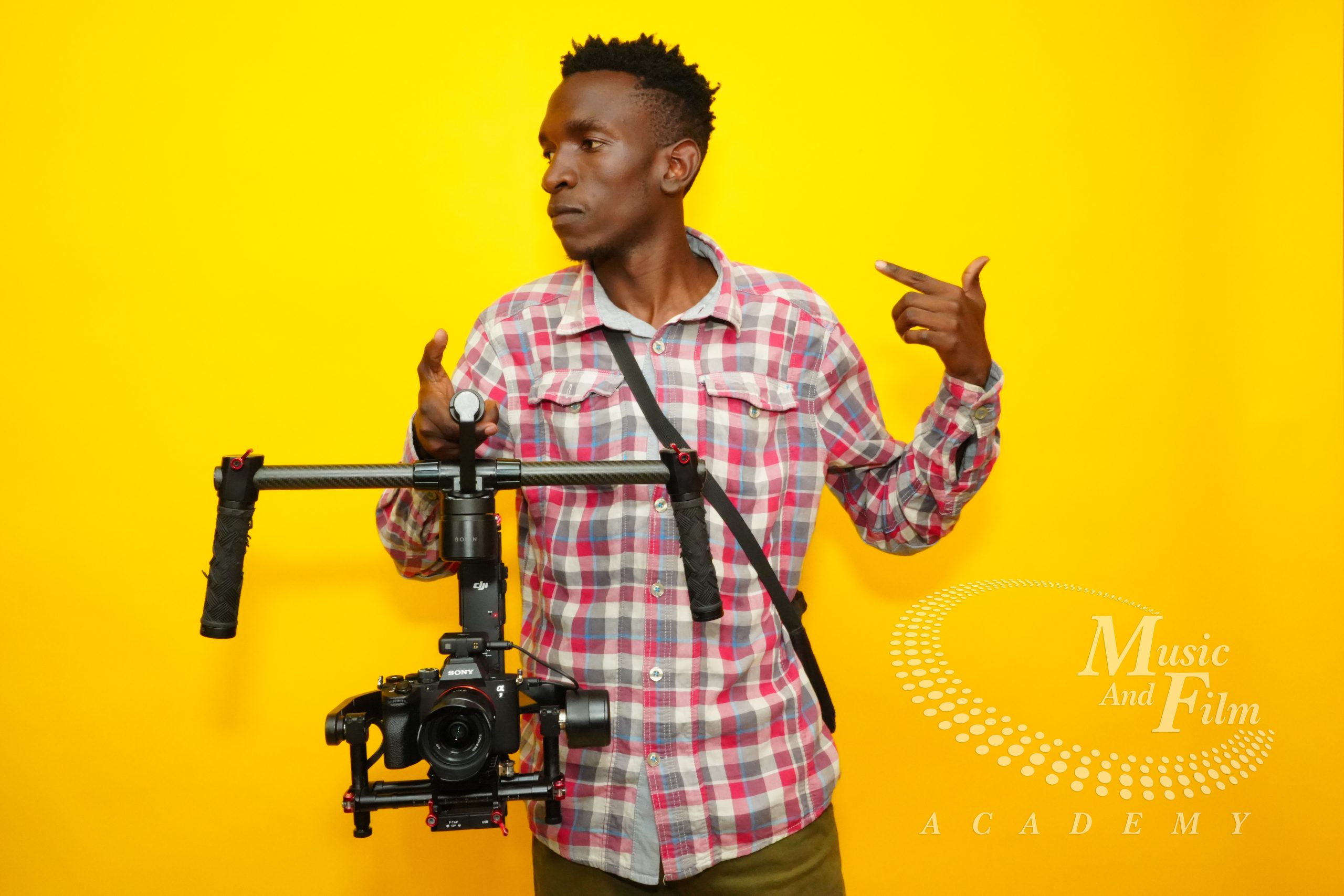 A student learning the skills for becoming a hybrid creator, operating a professional Sony mirrorless camera mounted on a dual-handle DJI Ronin gimbal stabilizer against a bright yellow background at the Music And Film Academy studio.