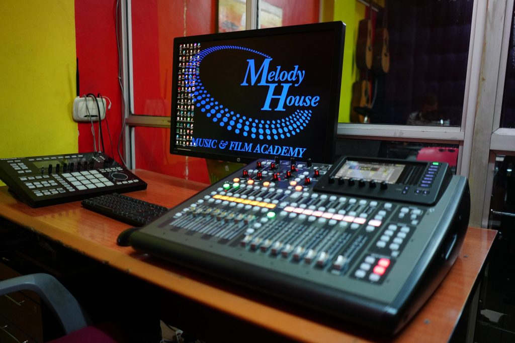 An enterprise digital mixing console with motorized faders and a routing display next to a pad controller inside a hybrid recording studio control room in Nairobi.