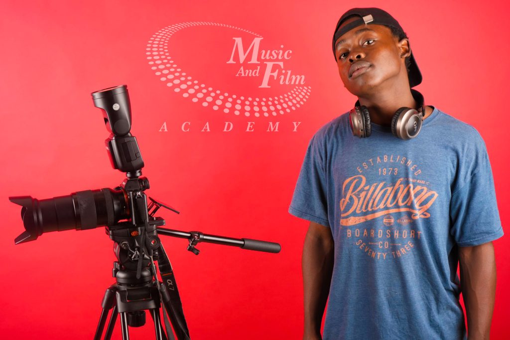 A Diploma in film production student wearing a blue Billabong t-shirt, over-ear headphones, and a backwards black cap poses next to a professional DSLR camera setup with an attached speedlight flash and fluid-head tripod against a red backdrop.