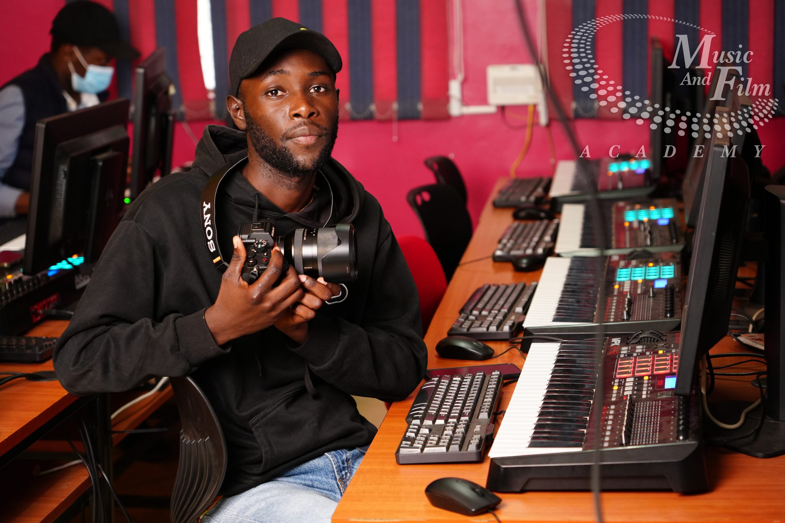 A digital media student holding a professional camera while sitting at a post-production workstation to structure a video editing workflow at Music and Film Academy.