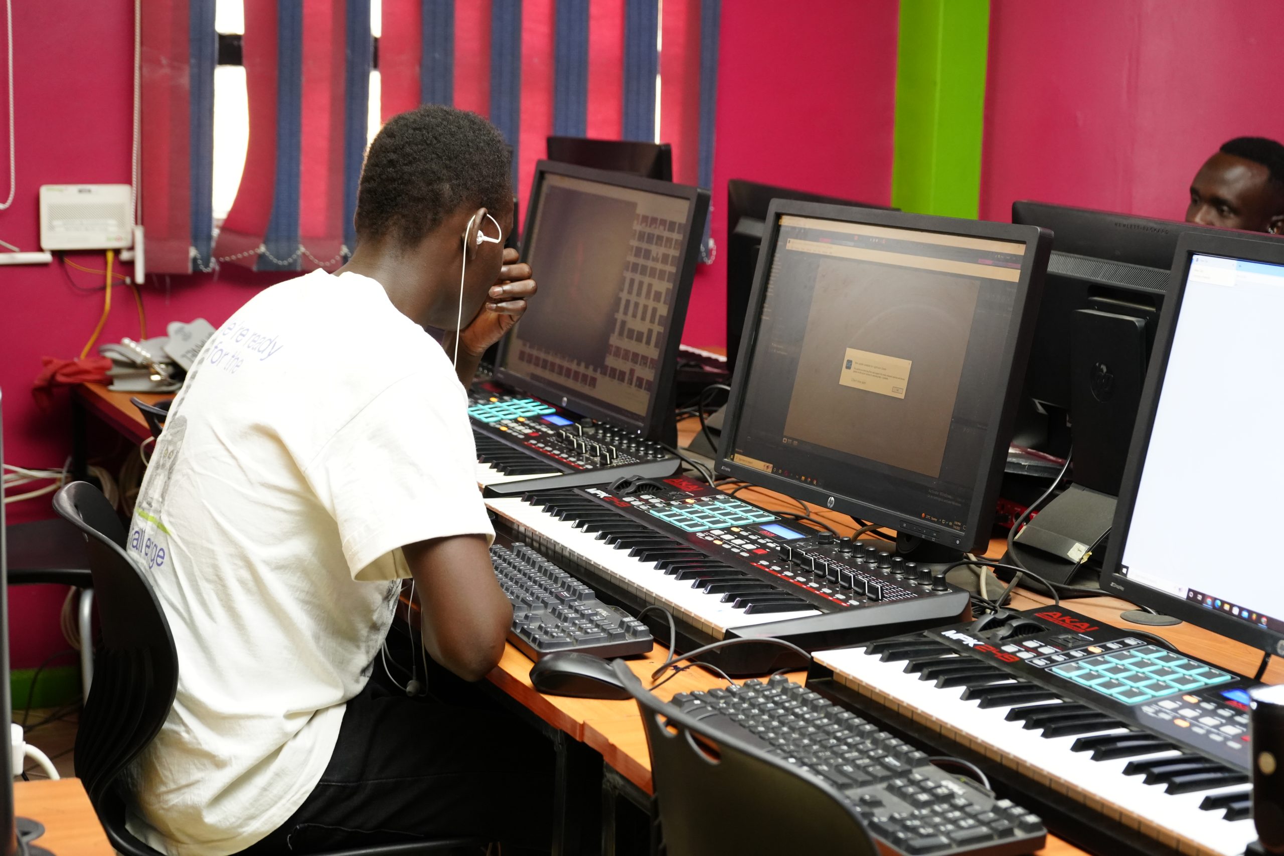 An audio student in a white t-shirt uses an independent digital workstation equipped with an Akai MPK249 MIDI keyboard controller, a computer monitor, and a keyboard during a practical class inside a music technology lab in Nairobi