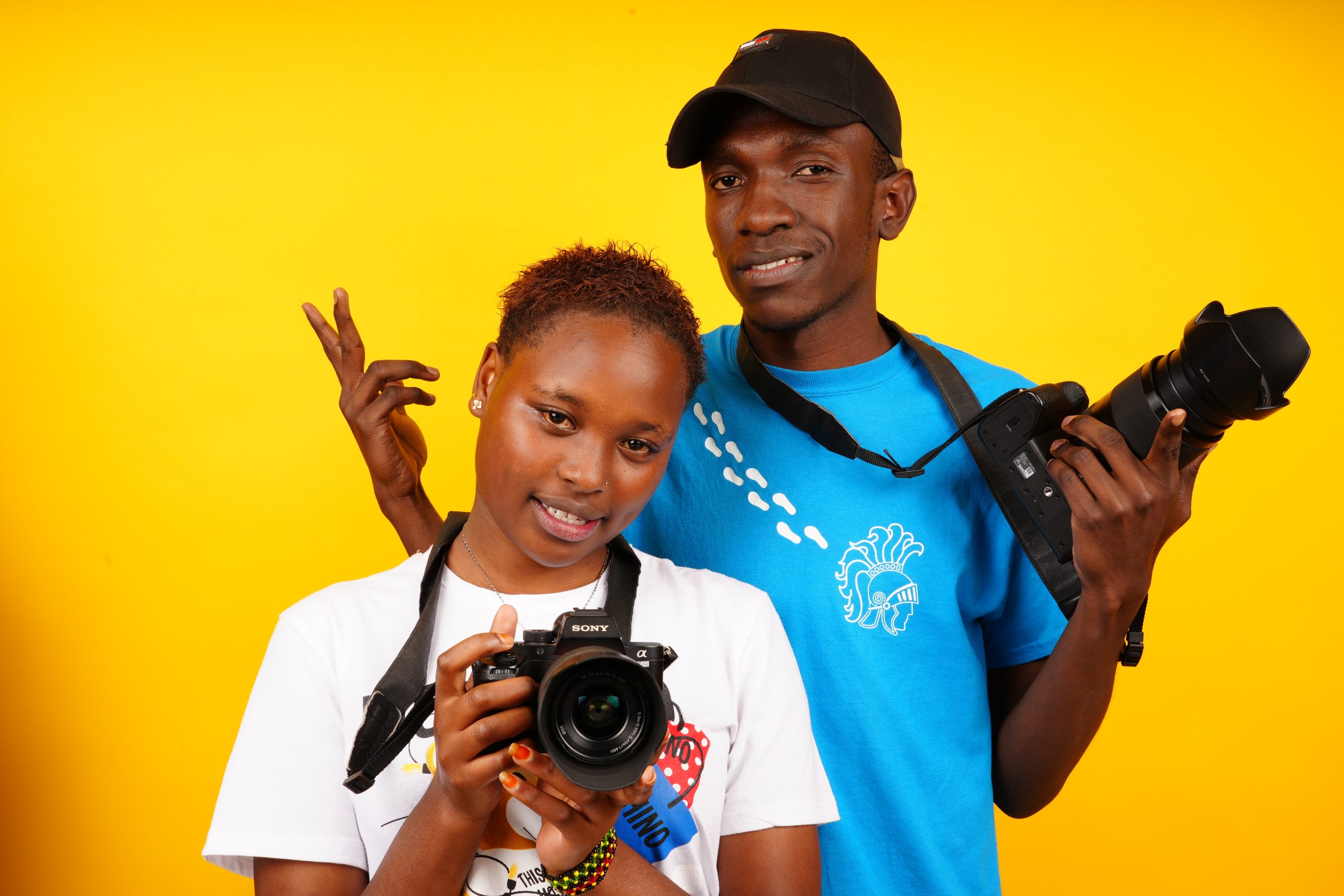 Two visual arts students holding professional cameras during a practical studio training session at one of the best photography schools in Nairobi, featuring a vibrant yellow background.